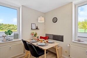 Ein Essbereich mit einem für zwei Personen gedecktem Tisch, einer Wanduhr und einem Fenster mit Blick auf Grünflächen.