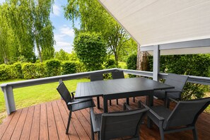 A covered outdoor deck with a table and chairs, surrounded by greenery and trees. - Wildgans 29 - Classic Scandinavian vacation home for up to 6 people (Mirow)