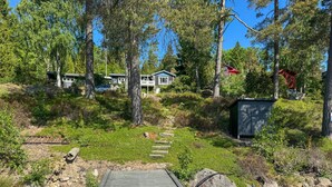 A wooden deck leading to a house surrounded by trees, with a small shed and a red structure in the background. - Cozy home in Motala with sauna (Motala)