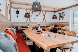 Ein gemütliches Interieur mit Holzbalken, einem langen Holztisch, Bänken und verschiedenen Wanddekorationen, darunter Tiergeweihe und hängende Objekte.