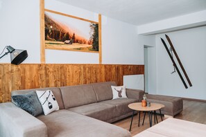 Ein modernes Wohnzimmer mit einer Wand in Holzoptik, einem großen gerahmten Bild, einer Couch mit Zierkissen, einem kleinen runden Tisch und Skiern, die an der Wand lehnen.