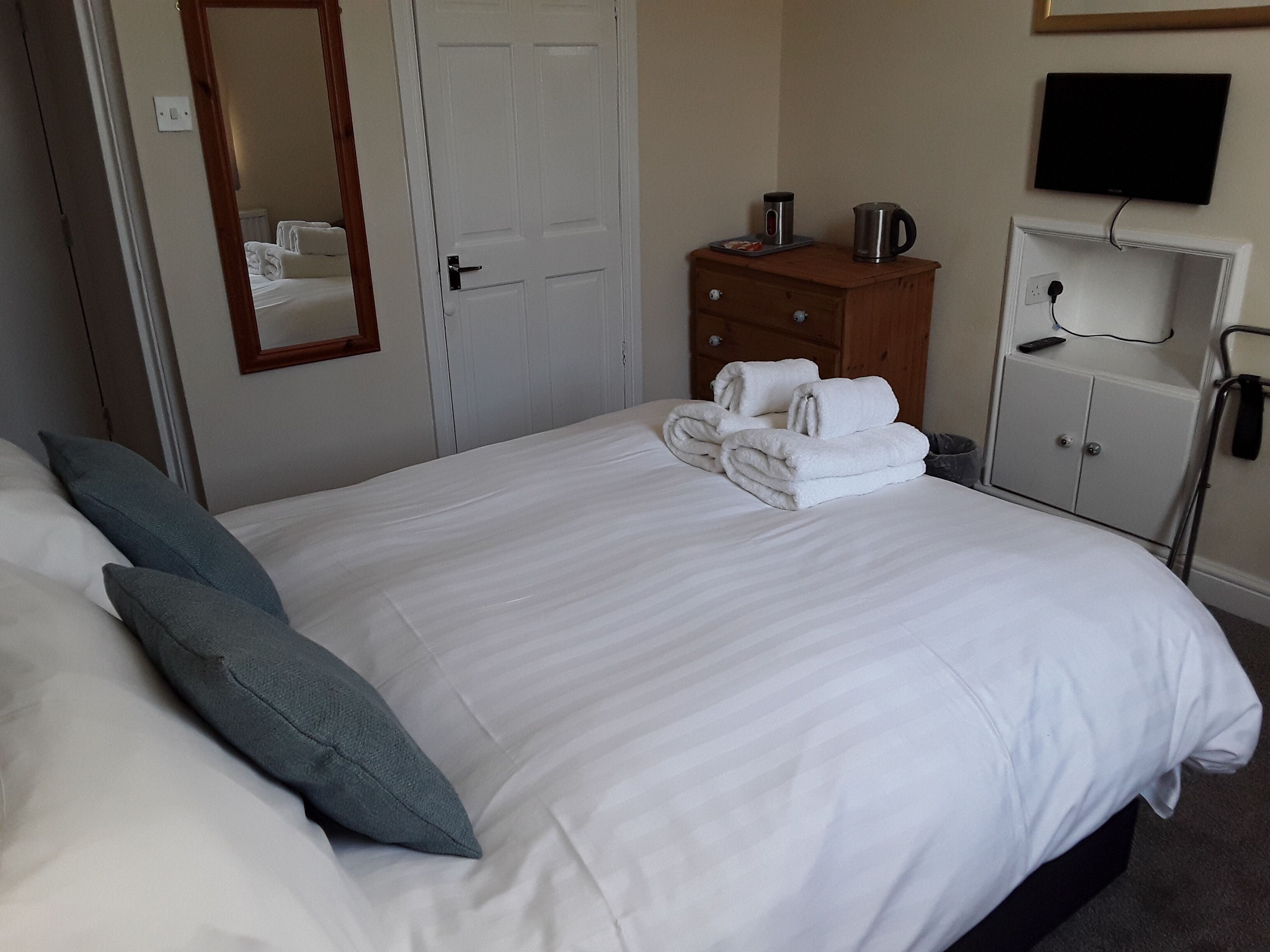 A neatly made bed with white linens and two pillows, a wooden dresser with a kettle and a cup, a mirror on the wall, and a television mounted above a cabinet.