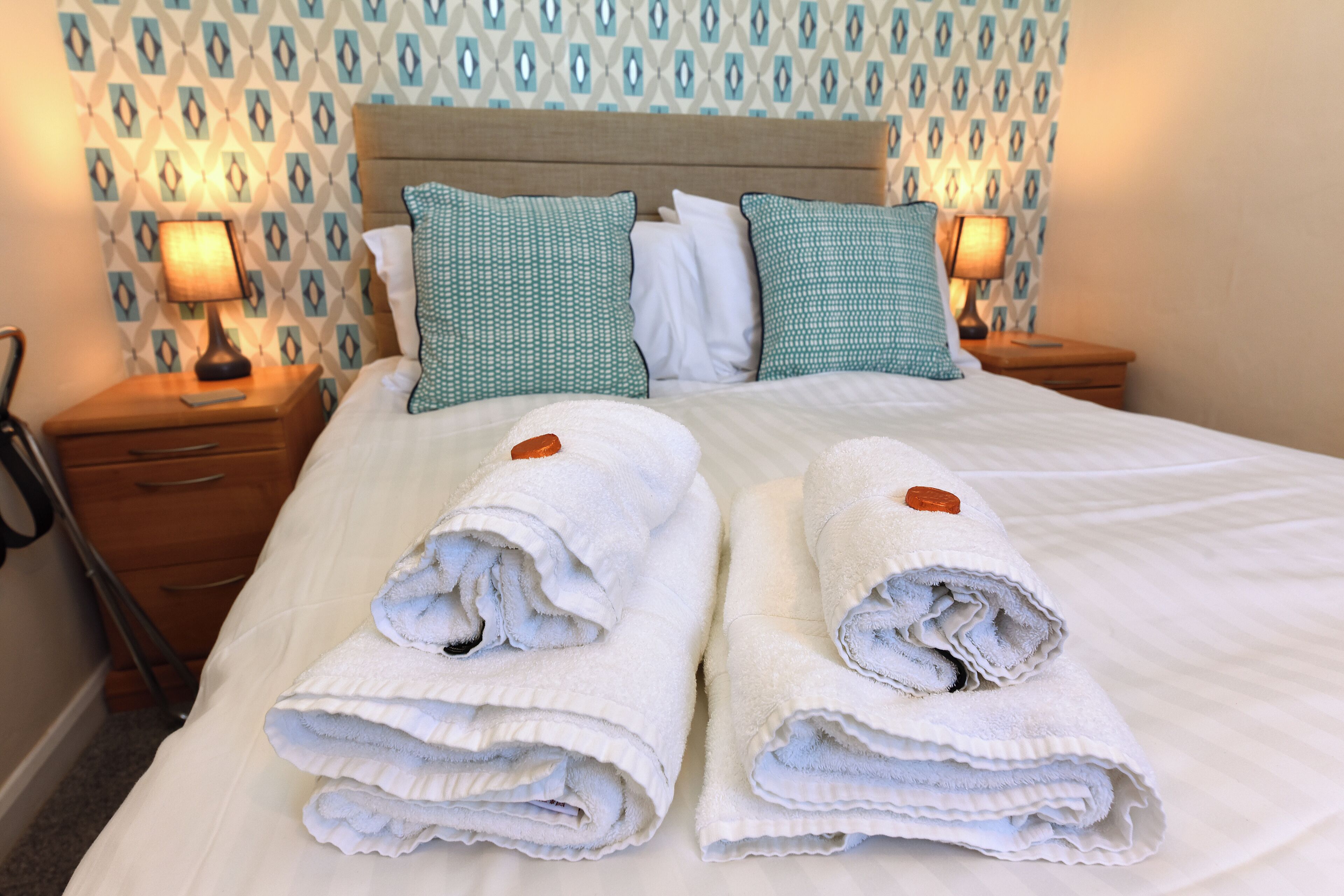 A neatly made bed with white linens and two rolled towels, a wooden nightstand with a lamp, and patterned wallpaper.