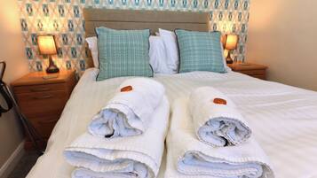A neatly made bed with white linens and two rolled towels, a wooden nightstand with a lamp, and patterned wallpaper.