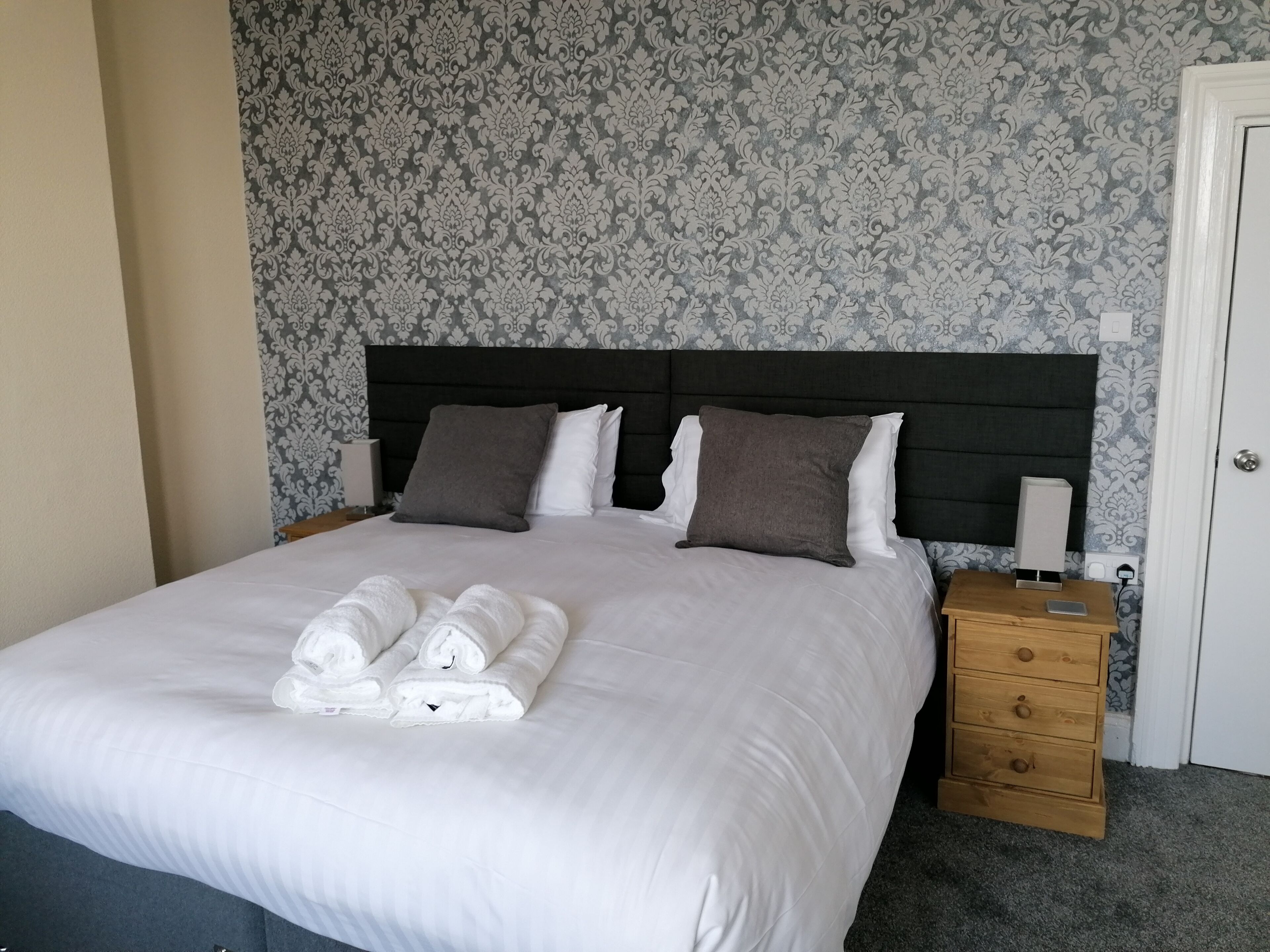 A neatly made bed with white linens and rolled towels, a dark headboard, a wooden nightstand with a lamp, and a patterned wallpaper.
