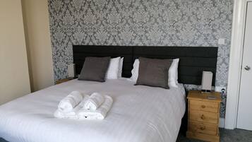 A neatly made bed with white linens and rolled towels, a dark headboard, a wooden nightstand with a lamp, and a patterned wallpaper.