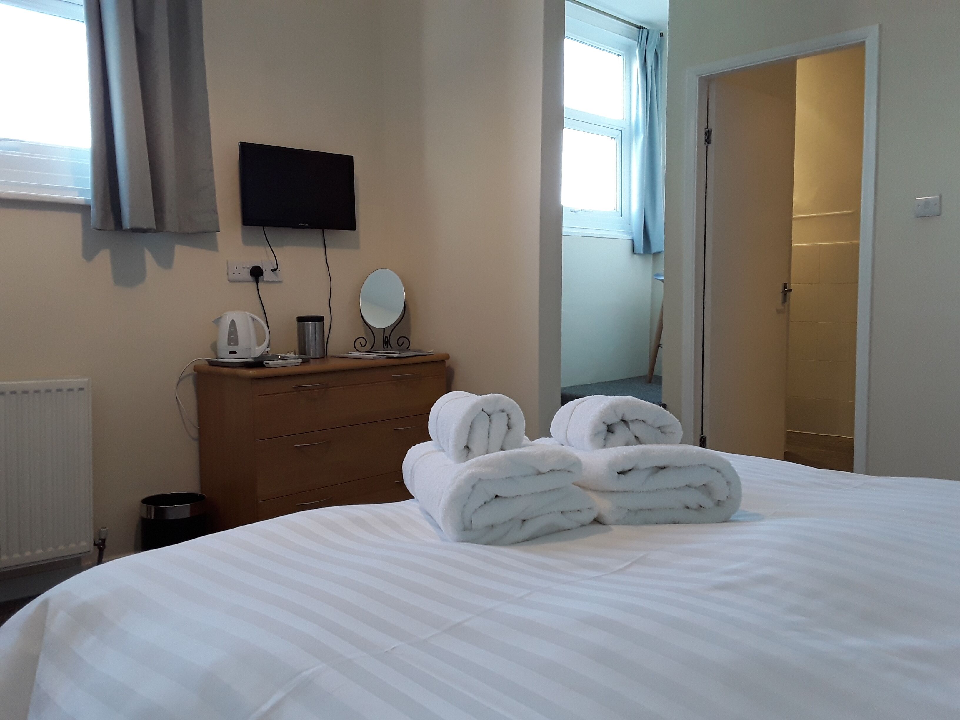 Habitación de hotel con cama, cómoda, televisor de pantalla plana, espejo y baño visible a través de una puerta abierta.