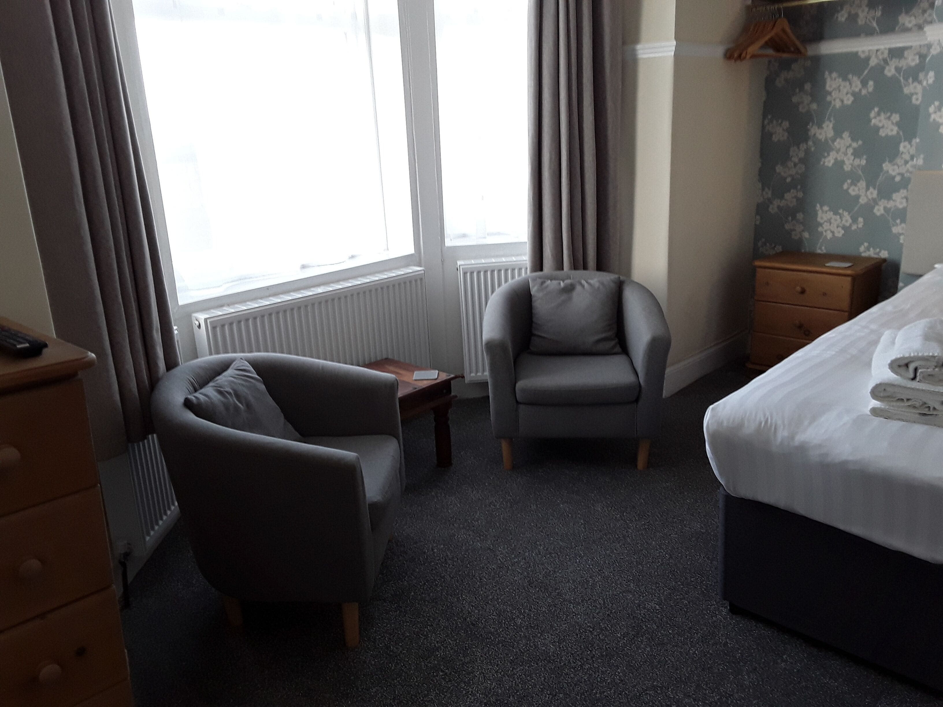 A hotel room with a bed, a grey armchair, a small table, a wooden dresser, and a radiator.