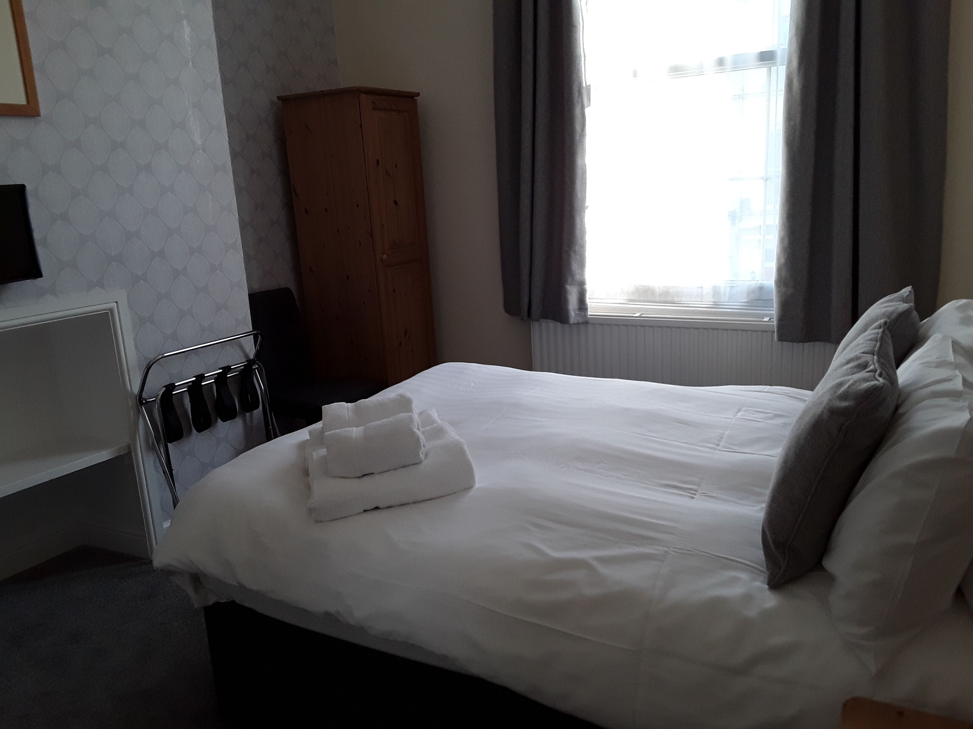 A hotel room with a bed, a wooden wardrobe, a radiator, and a window with curtains.