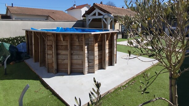 A wooden structure with a blue roof, possibly a shed or playhouse, in a backyard with a lawn and a tree.