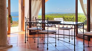 Une pièce avec une table et des chaises, de grandes fenêtres avec des rideaux, et une vue sur la verdure et les montagnes.
