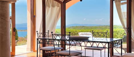 A room with a table and chairs, large windows with curtains, and a view of greenery and mountains.