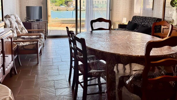A room with a dining table, chairs, a sofa, a TV, and a view of the sea through large windows. - THE TAMARISSIERE RETREAT (Agde)