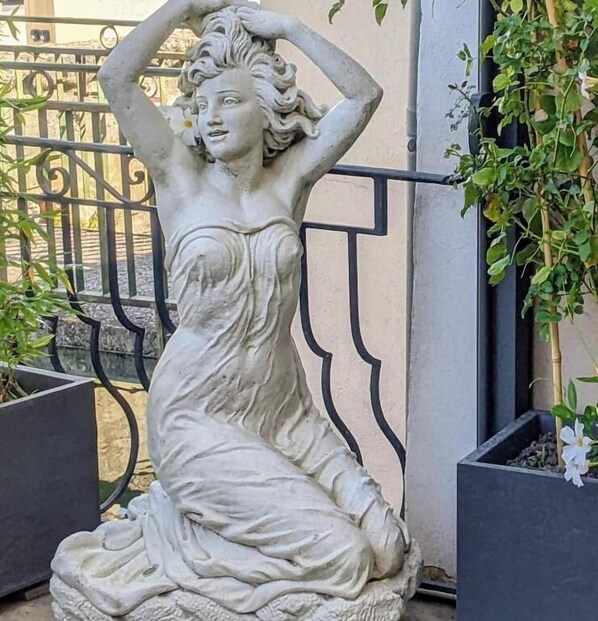 Uma estátua de mulher num vestido, ao lado de uma grade de metal e vasos com plantas.