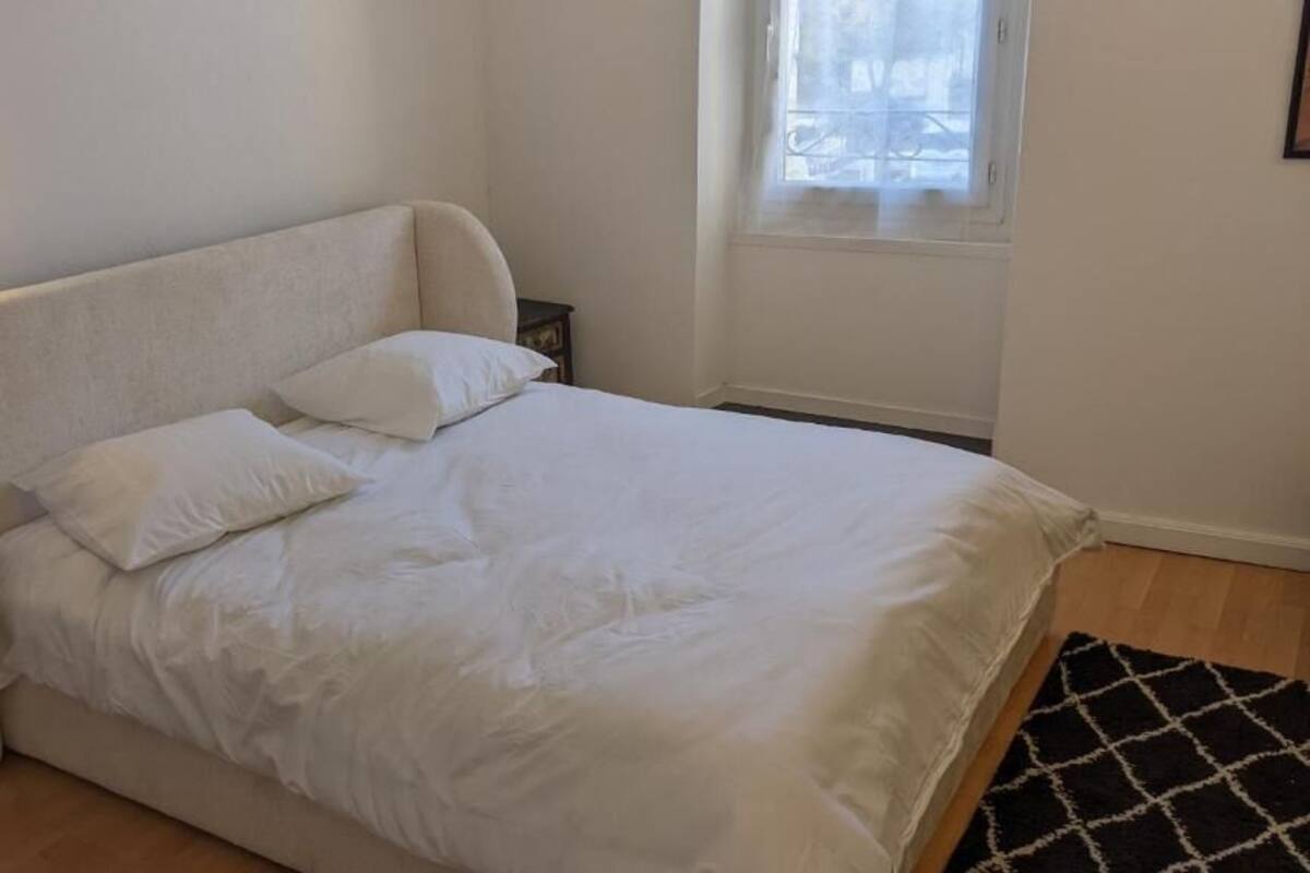 Una cama con ropa de cama blanca, una lámpara de noche, una mesita auxiliar pequeña y una ventana con vistas.