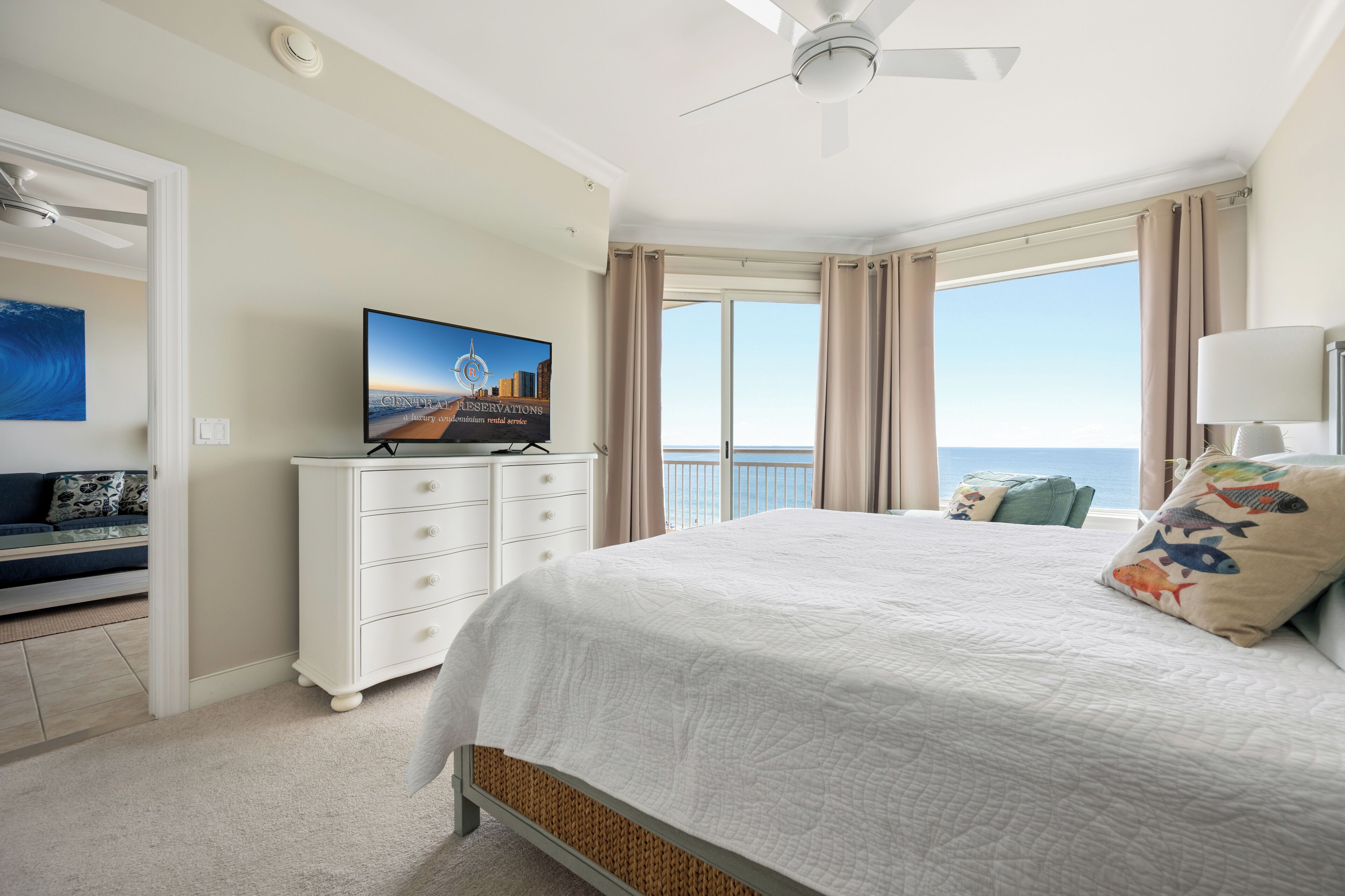 A bedroom with a bed, a television on a dresser, a ceiling fan, and a view of the ocean.