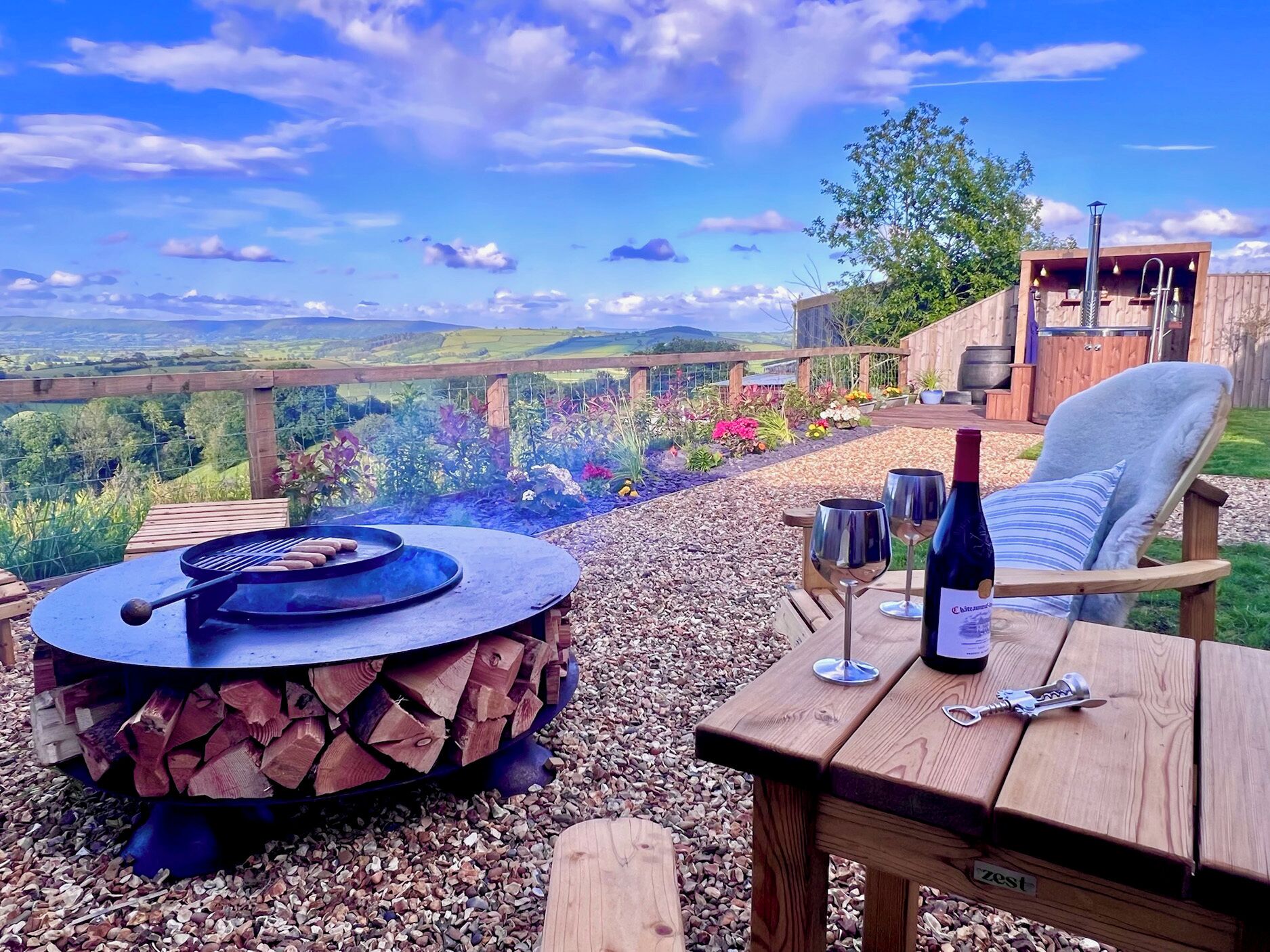 Une terrasse extérieure avec un foyer, une table avec des verres à vin et une bouteille, et une vue sur la campagne.