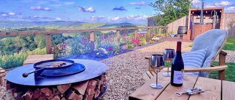 An outdoor patio with a fire pit, a table with wine glasses and a bottle, and a view of the countryside.