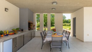 A modern outdoor kitchen with a dining area, large windows, and a view of greenery. - Villa Bateli by Villas Guide (Barban)