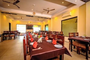 A dining area with multiple tables set for a meal, ceiling fans, recessed lighting, and a large window providing natural light. - CHIRAIYA RESORT AND HOTELS (Uttarakhand)