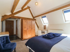A bedroom with a sloped ceiling, wooden beams, a wardrobe, a desk, a chair, and a bed with a blue blanket. - Cefnbron (Caernarfon)