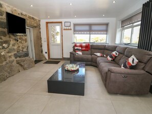 A living room with a stone wall, a flat-screen TV, a sectional sofa, and a coffee table. - Cefnbron (Caernarfon)