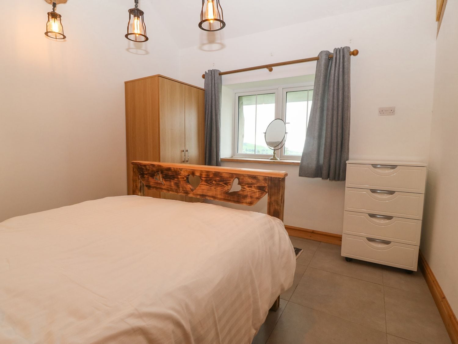 A bedroom with a bed, a wooden headboard, a dresser, a mirror, and a window with curtains.