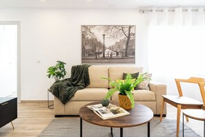 Una sala de estar con un sofá, una mesa de centro, una planta, un cuadro enmarcado y una silla.