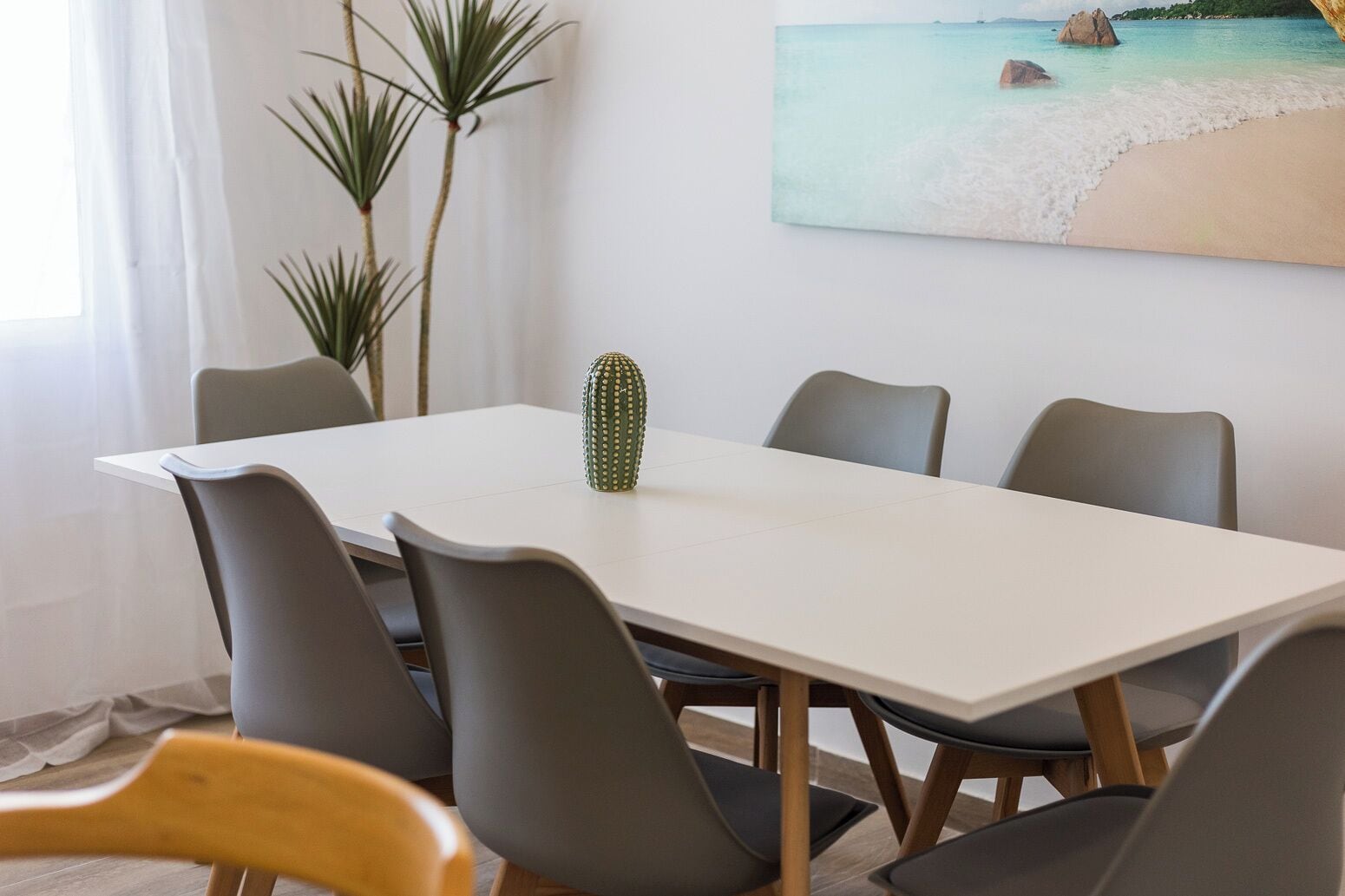 Un coin repas avec une table blanche, six chaises grises, un cactus en guise de centre de table et un tableau représentant une scène de plage accroché au mur.