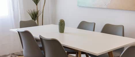 Un comedor con una mesa blanca, seis sillas grises, un centro de mesa con un cactus y un cuadro de una playa en la pared.