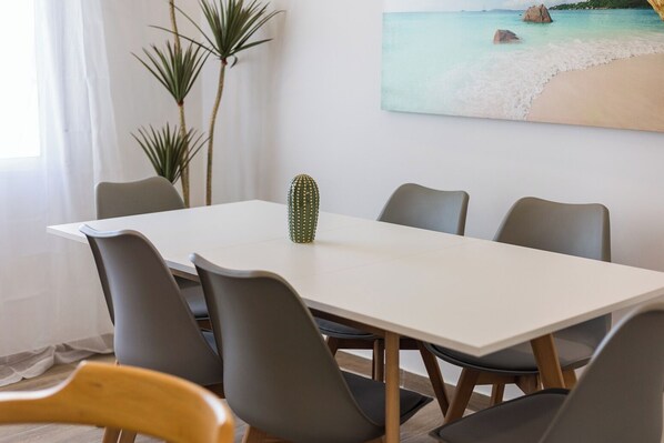Un comedor con una mesa blanca, seis sillas grises, un centro de mesa con un cactus y un cuadro de una playa en la pared.