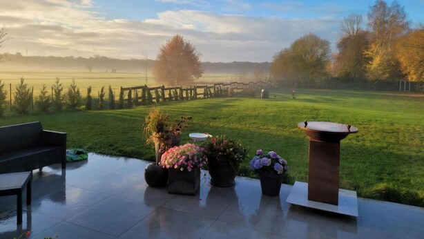 A patio with a view of a grassy field, a wooden fence, and a large tree.