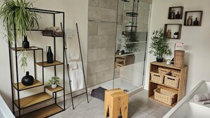 A modern bathroom with a glass shower enclosure, wooden shelves with plants, and a small wooden stool.