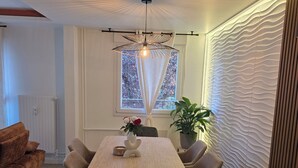 A modern dining area with a wooden table, grey chairs, a hanging light fixture, a radiator, a window with curtains, and a potted plant. - Luxury Apartment • 3 Suites • 2 Parking Lots • 5 min Downtown (Strasbourg)