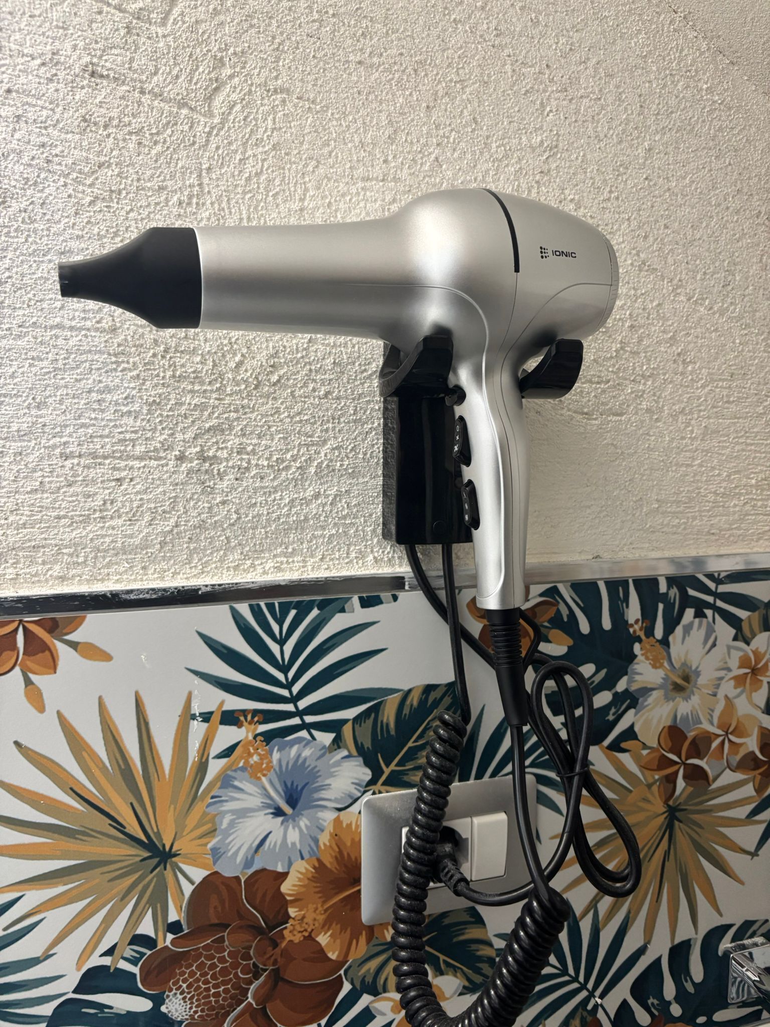 Un asciugacapelli fissato al muro, con carta da parati a motivi floreali.