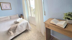 A bedroom with a bed, a bedside table with a book, a window with curtains, and a wall-mounted light switch.