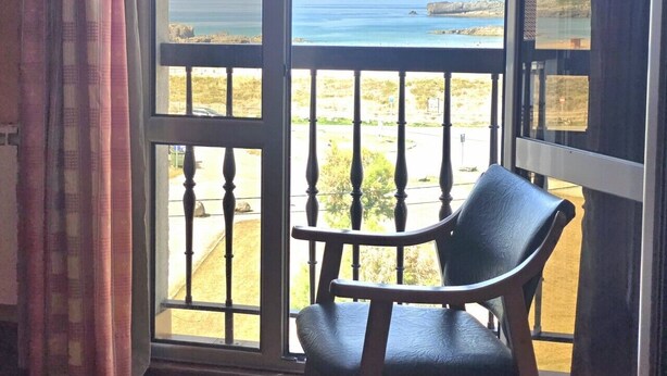 A balcony with a view of the sea, a chair, and a window with curtains.