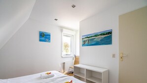 Quarto com duas camas, um banco branco, uma pintura de uma praia e uma janela com vista para o verde.