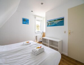 A bedroom with two beds, a white bench, a painting of a beach, and a window with a view of greenery. - Home With Dune Views (Vlissingen)