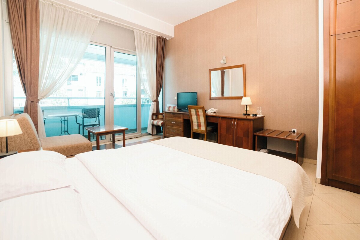Habitación de hotel con dos camas, un escritorio con computadora, una silla, una mesita y un balcón con sillón y mesa.