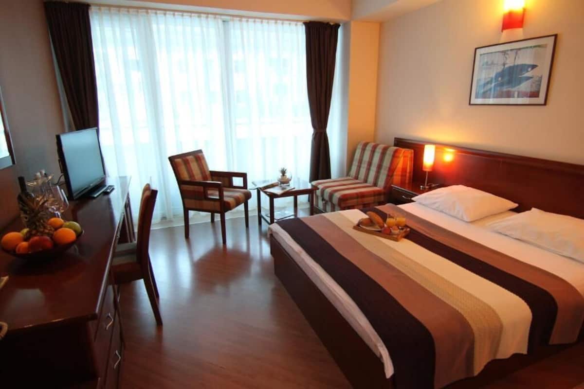 A hotel room with a bed, a desk with a computer, a chair, a small table with a lamp, and a window with curtains.