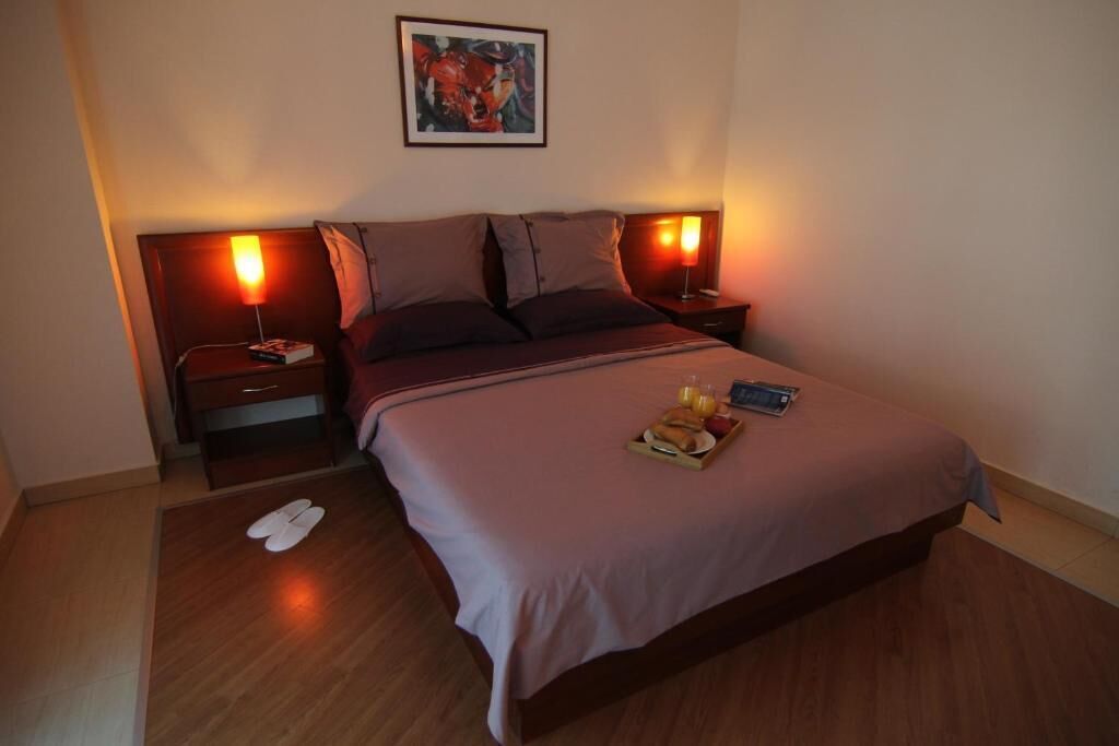 A hotel room with a bed, two bedside tables, a nightstand, a framed picture on the wall, and a pair of slippers on the floor.