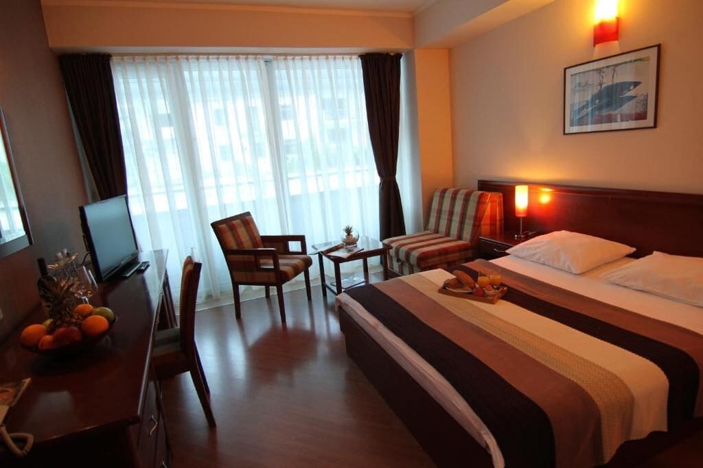 A hotel room with a bed, a desk with a computer, a chair, a small table, and a window with curtains.