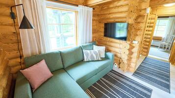 En stuga med träpanel på insidan, en grön soffa, en tv som är monterad på väggen och ett fönster med gardiner.