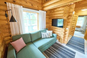A log cabin interior with a green sofa, a TV mounted on the wall, and a window with curtains.