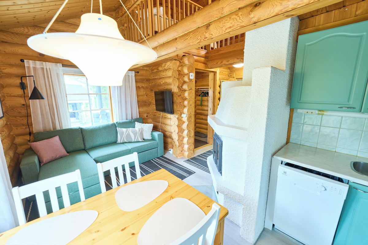 A log cabin interior with a wooden dining table, a green sofa, a kitchen area with a white dishwasher, and a wooden staircase.