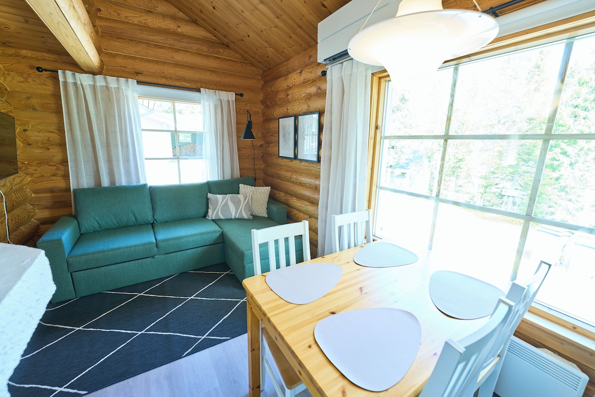 A wooden cabin interior with a green sofa, a dining table with chairs, and a large window.