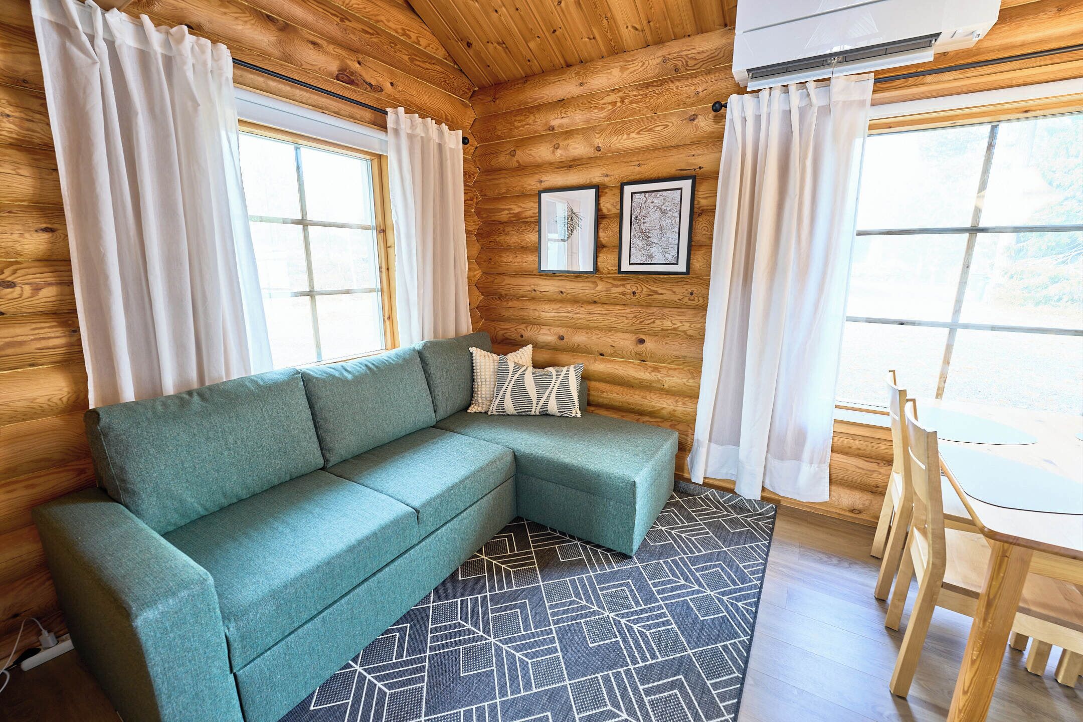 Interior de una cabaña de madera con un sofá verde, una mesa de comedor y dos cuadros en la pared.