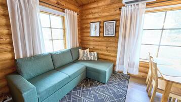 Interior de una cabaña de madera con un sofá verde, una mesa de comedor y dos cuadros en la pared.