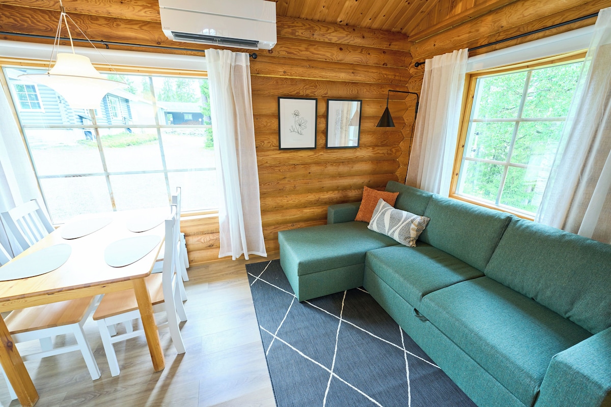 A wooden cabin interior with a dining table, green sofa, and framed pictures on the wall.
